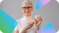 A smiling woman with gray hair and glasses holds up a jar of cosmetic cream.