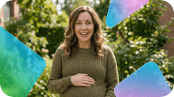 A smiling pregnant woman in a green sweater holds her belly, surrounded by trees.